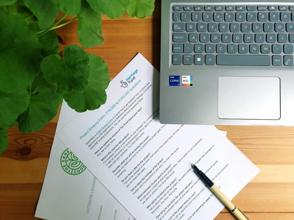 Photo of a desk showing part of a plant, laptop, pen and grant application forms