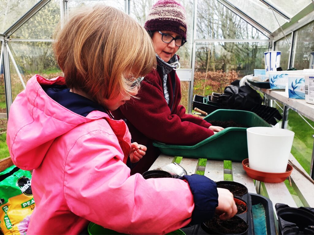 Photo of a young girl in a pink coat and glasses and an older woman in a maroon coat, glasses and beanie hat potting seedlings in a greenhouse